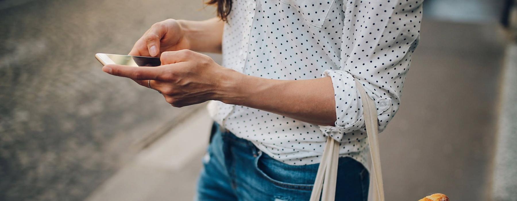 woman walks down the street and texts with a bag of groceries on her arm
